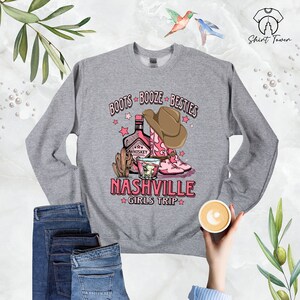 May include: Gray sweatshirt with a pink and brown graphic design that says "Boots Booze Besties Nashville Girls Trip". The graphic includes a bottle of whiskey, a shot glass, and cowboy boots.