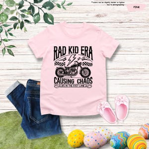 May include: Pink t-shirt with a black and white graphic of a motorcycle with the text "Rad Kid Era Causing Chaos Life in the Fast Lane"