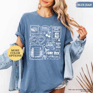 May include: Blue jean colored t-shirt with white graphic illustrations of books, hearts, and text. The shirt includes the phrases "Grumpy x Sunshine", "Just One More Chapter", "Born to Read", and "One Bed at the Inn".