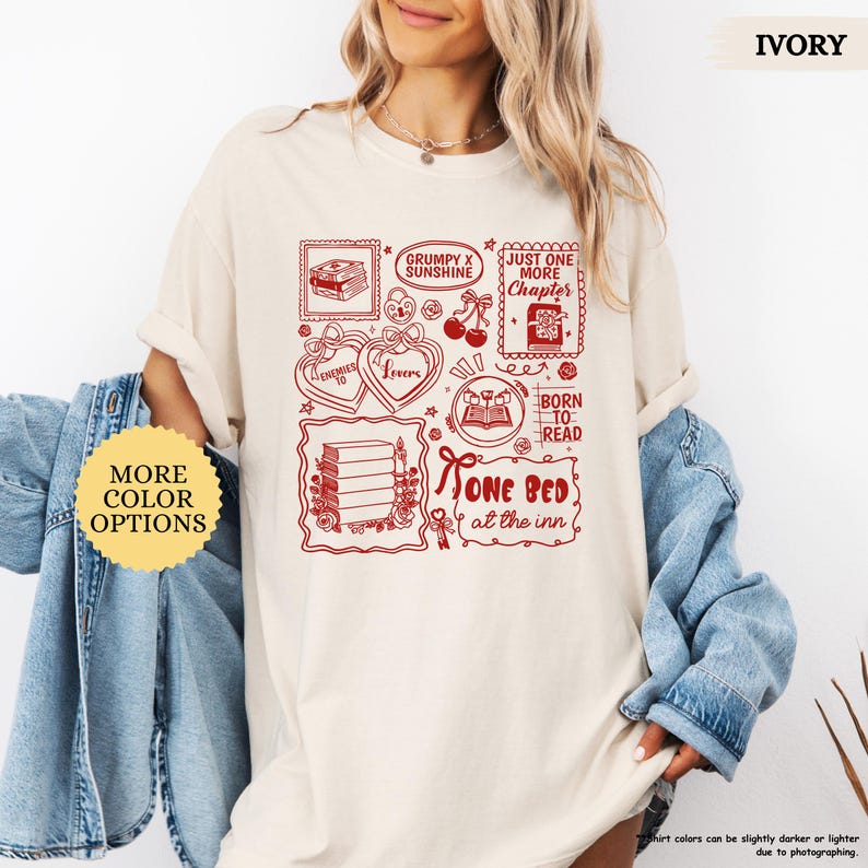 May include: Ivory-colored t-shirt with a red graphic featuring book-themed illustrations and text. The design includes stamps with books, hearts, cherries, and the phrases "Grumpy x Sunshine," "Just One More Chapter," and "One Bed at the Inn."