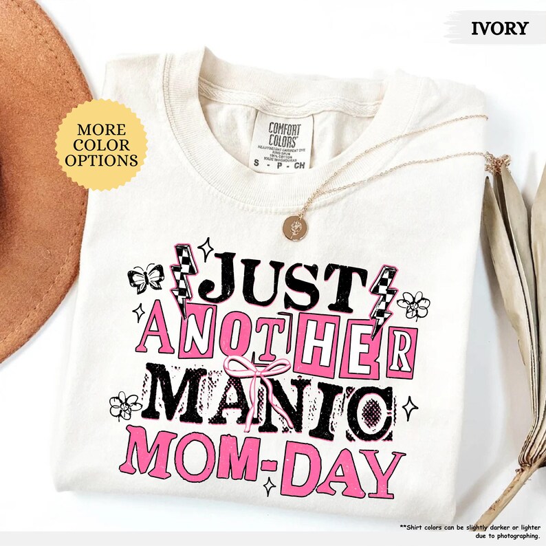 May include: Ivory-colored t-shirt with the text "JUST ANOTHER MANIC MOM-DAY" in black and pink. The shirt features a crew neck and a small gold necklace. The design includes lightning bolts, flowers, and a bow.