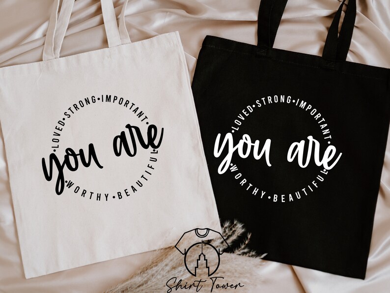 K&ouml;nnte beinhalten: Zwei Einkaufstaschen mit dem Text "you are" in einem Kreis, umgeben von den W&ouml;rtern "loved, strong, important, worthy, beautiful". Eine Tasche ist wei&szlig; mit schwarzem Text und die andere ist schwarz mit wei&szlig;em Text.