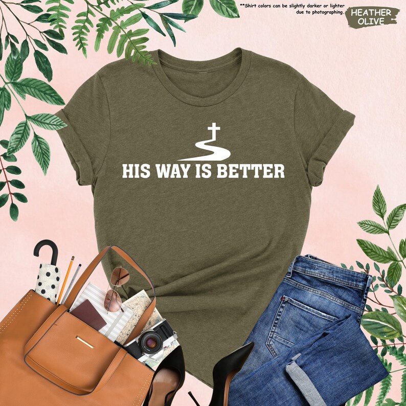 May include: Heather olive green t-shirt with the text "HIS WAY IS BETTER" in white. The shirt features a graphic of a cross above a winding path. The shirt is paired with blue jeans, a brown purse, and black heels.