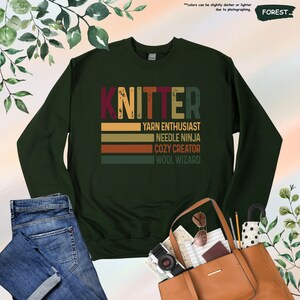 May include: A dark green sweatshirt with the word "KNITTER" in large, retro-style letters. Below the word are five horizontal stripes in shades of brown, orange, and yellow. The text "YARN ENTHUSIAST", "NEEDLE NINJA", "COZY CREATOR", and "WOOL WIZARD" is printed in white below the stripes.