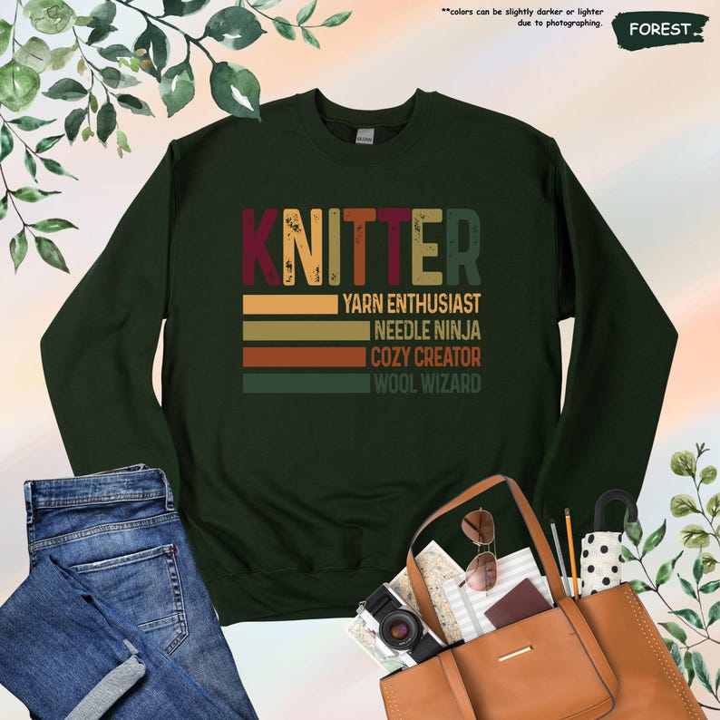 May include: A dark green sweatshirt with the word "KNITTER" in large, retro-style letters. Below the word are five horizontal stripes in shades of brown, orange, and yellow. The text "YARN ENTHUSIAST", "NEEDLE NINJA", "COZY CREATOR", and "WOOL WIZARD" is printed in white below the stripes.
