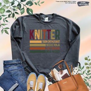 May include: Dark heather grey sweatshirt with the text "KNITTER" in a vintage style font. The text is layered over a series of colorful stripes. Below the text are the words "YARN ENTHUSIAST", "NEEDLE NINJA", "COZY CREATOR", and "WOOL WIZARD".