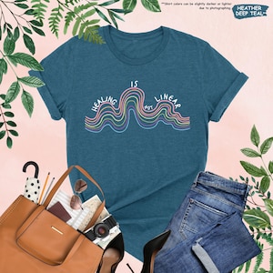 Op de afbeelding: Een diep-tealkleurig t-shirt met de tekst "HEALING IS NOT LINEAR" in een golvend ontwerp met pastelkleuren. Het shirt is gestyled met een blauwe jeans, een bruine totebag en zwarte hakken.