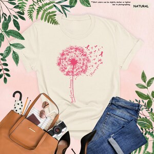 Puede incluir: Camiseta color crema con un dise&ntilde;o de diente de le&oacute;n rosa y las palabras "Breast Cancer Awareness" y "Never give up." La imagen incluye vaqueros azules, un bolso marr&oacute;n y tacones negros.