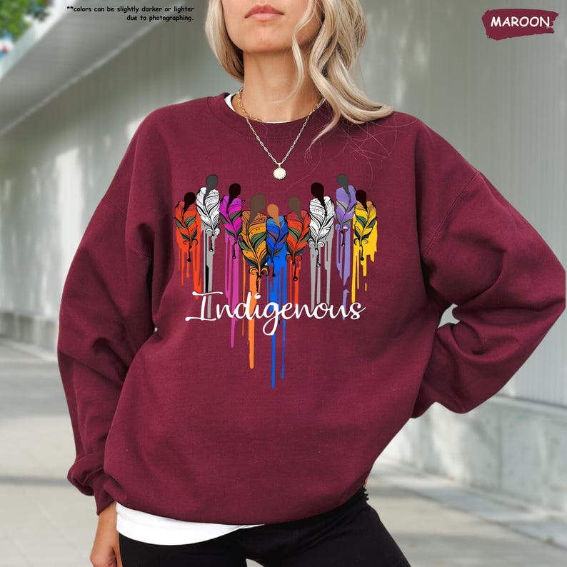 May include: A maroon sweatshirt with a graphic design of colorful figures with feathers and the word "Indigenous" in a dripping paint style.