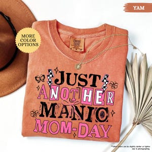 May include: A yam-colored t-shirt with the text "JUST ANOTHER MANIC MOM-DAY" in black and pink. The shirt has a crew neck and short sleeves. A gold necklace with a pendant is draped across the shirt. The words "MORE COLOR OPTIONS" are in a yellow circle.