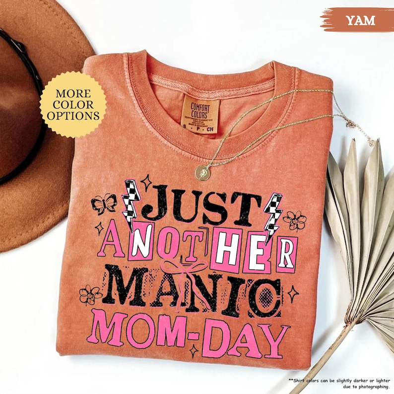 May include: A yam-colored t-shirt with the text "JUST ANOTHER MANIC MOM-DAY" in black and pink. The shirt has a crew neck and short sleeves. A gold necklace with a pendant is draped across the shirt. The words "MORE COLOR OPTIONS" are in a yellow circle.