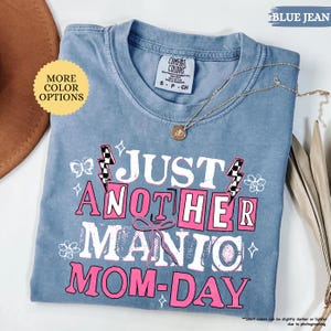 May include: Blue jean colored t-shirt with the text "JUST ANOTHER MANIC MOM-DAY" in white and pink. The shirt has a crew neck and short sleeves. A gold necklace with a pendant is draped across the shirt.