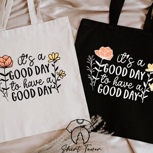 May include: Two tote bags with the text "it's a GOOD DAY to have a GOOD DAY" printed on them. The bags are made of canvas and have floral designs. One bag is white and the other is black.