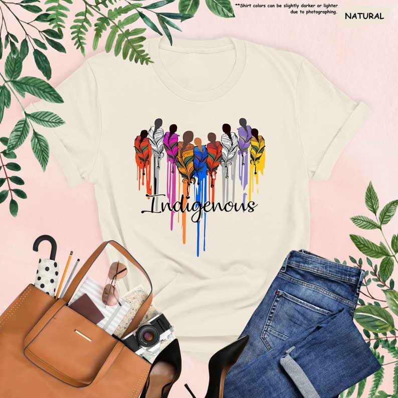 May include: A beige t-shirt with a graphic of a group of people with colorful feathers and the word "Indigenous" in a dripping paint style.
