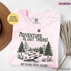 May include: A light pink t-shirt with the text "ADVENTURE IS OUT THERE! BUT SO ARE SERIAL KILLERS" and a forest graphic. The shirt has a crew neck and short sleeves. A gold necklace with a pendant is on the shirt.