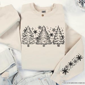 May include: A cream-colored sweatshirt with a black outline of a row of Christmas trees and snowflakes. The sweatshirt has a black snowflake design on the sleeve.