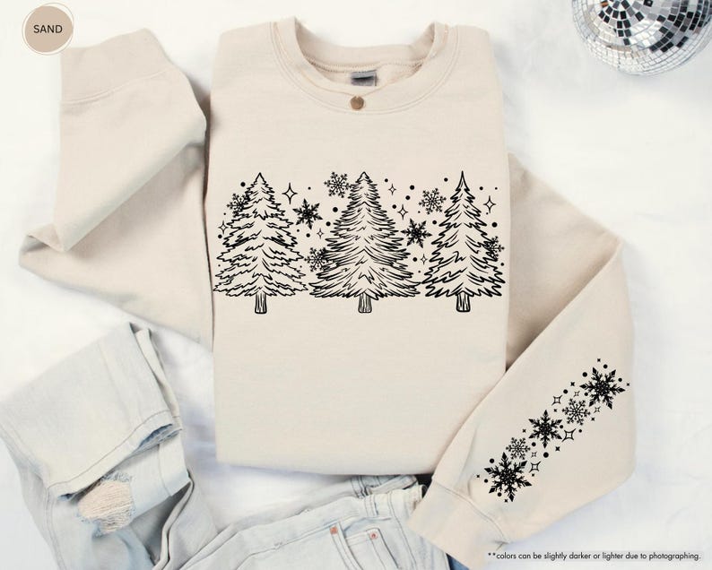 May include: A cream-colored sweatshirt with a black outline of a row of Christmas trees and snowflakes. The sweatshirt has a black snowflake design on the sleeve.