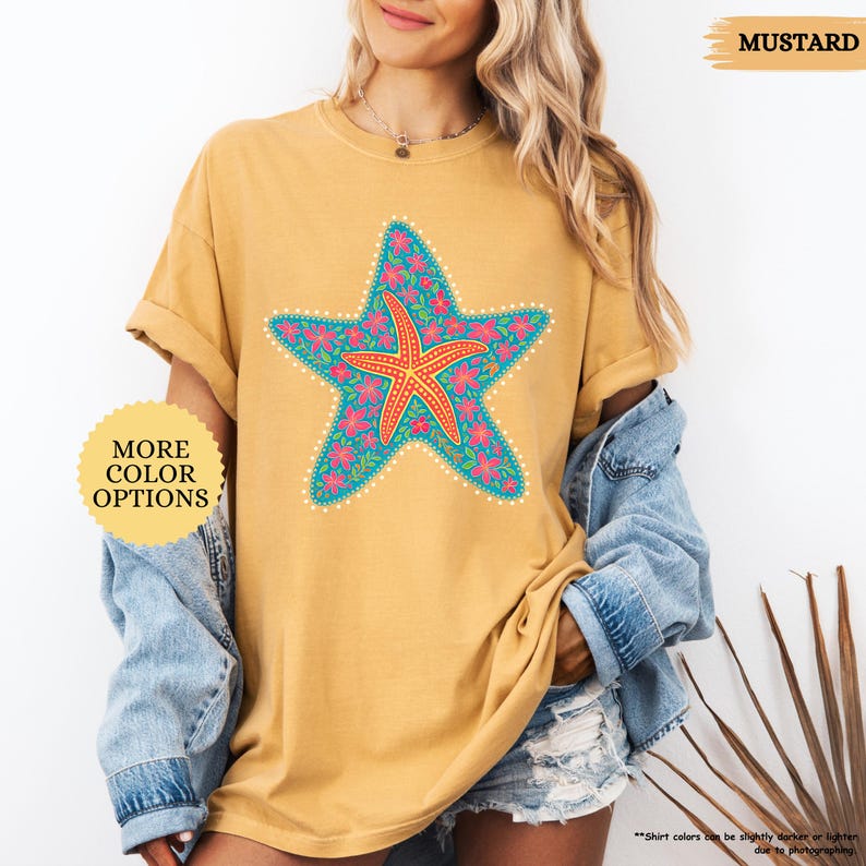 May include: Mustard-colored t-shirt featuring a large, detailed starfish design. The starfish is a vibrant mix of teal, pink, and orange, with a floral pattern. The shirt has the text "MORE COLOR OPTIONS" and "MUSTARD".