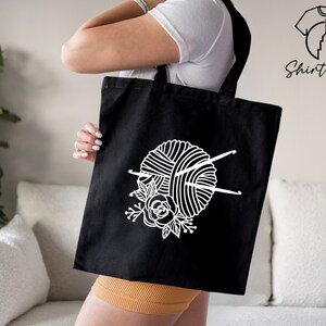 Pu&ograve; includere: Una borsa tote nera con un designo bianco di un gomitolo di lana, ferri da maglia e fiori.
