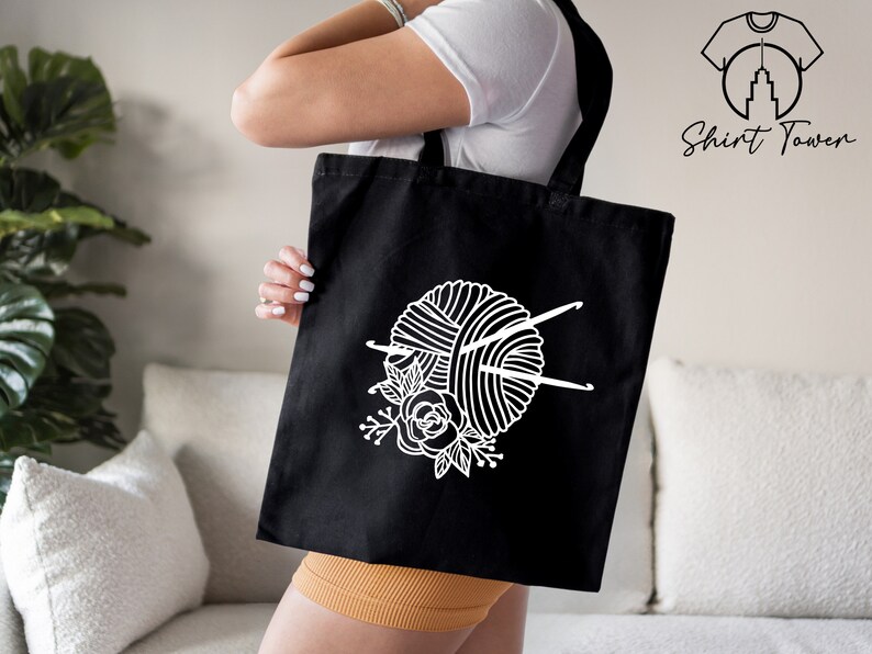 Pu&ograve; includere: Una borsa tote nera con un designo bianco di un gomitolo di lana, ferri da maglia e fiori.