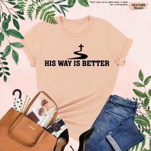 May include: Heather peach t-shirt with the text "HIS WAY IS BETTER" in black. The shirt features a cross design above the text. The image also includes a brown purse, blue jeans, and black high heels.
