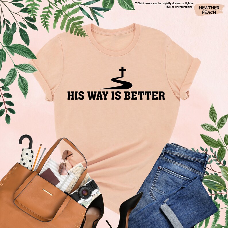 May include: Heather peach t-shirt with the text "HIS WAY IS BETTER" in black. The shirt features a cross design above the text. The image also includes a brown purse, blue jeans, and black high heels.