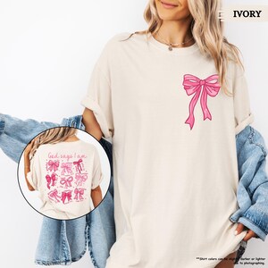 K&ouml;nnte beinhalten: Ein wei&szlig;es T-Shirt mit einer rosa Schleife auf der Vorderseite. Die R&uuml;ckseite des Shirts tr&auml;gt den Text "God says I am" und ein Raster aus rosa Schleifen.