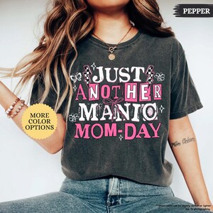May include: Dark gray t-shirt with the text "JUST ANOTHER MANIC MOM-DAY" in white and pink. The shirt has a vintage, worn look. The words are arranged in a playful, eye-catching design. The shirt also has the word "PEPPER" on the top right.