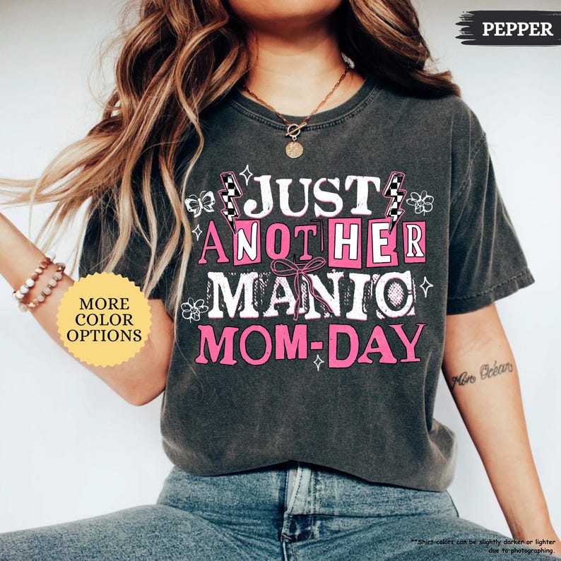 May include: Dark gray t-shirt with the text "JUST ANOTHER MANIC MOM-DAY" in white and pink. The shirt has a vintage, worn look. The words are arranged in a playful, eye-catching design. The shirt also has the word "PEPPER" on the top right.
