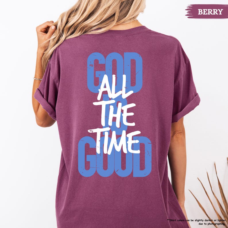 May include: Berry-colored t-shirt with the words "GOD ALL THE TIME GOOD" in blue and white lettering. The shirt has rolled-up sleeves and a relaxed fit. The text is distressed, giving it a vintage look.