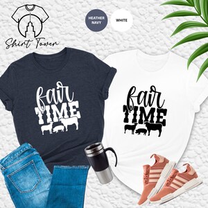 May include: Two t-shirts with the text "fair time" in white lettering and a row of farm animals below. The shirts are heather navy and white. The heather navy shirt is on a pair of blue jeans and a black travel mug.