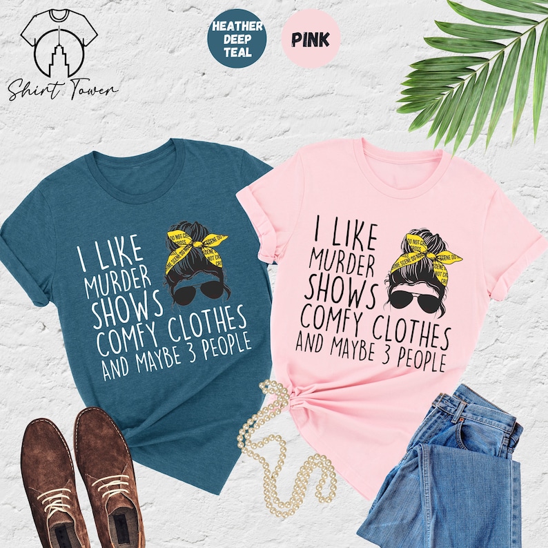May include: Two t-shirts with a graphic of a woman with a messy bun wearing sunglasses and a yellow crime scene tape headband. The text on the shirts reads "I like murder shows comfy clothes and maybe 3 people". One shirt is heather deep teal and the other is pink.