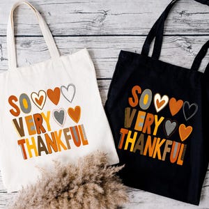 Pode incluir: Duas sacolas de tecido com o texto "So Very Thankful" em letras coloridas e cora&ccedil;&otilde;es. Uma sacola &eacute; de lona natural e a outra &eacute; preta.