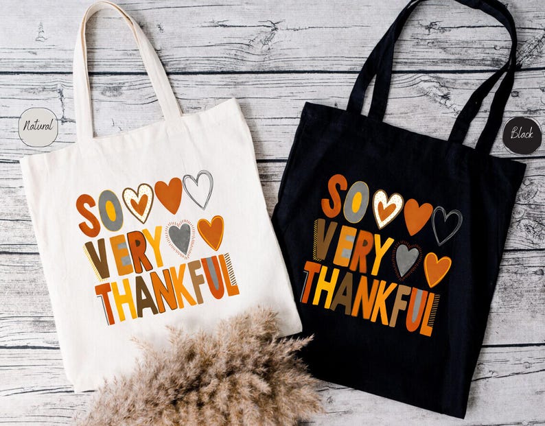 Pode incluir: Duas sacolas de tecido com o texto "So Very Thankful" em letras coloridas e cora&ccedil;&otilde;es. Uma sacola &eacute; de lona natural e a outra &eacute; preta.