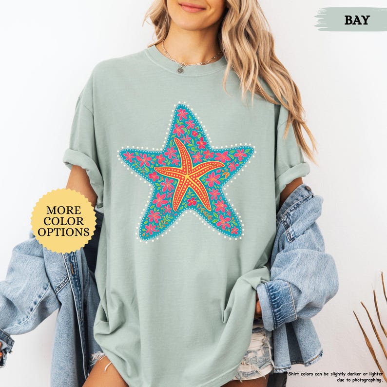 May include: A light green t-shirt featuring a large, colorful starfish design. The starfish is orange and surrounded by pink flowers and a blue border. The shirt has a relaxed fit and short sleeves. The text "MORE COLOR OPTIONS" is in a yellow circle.