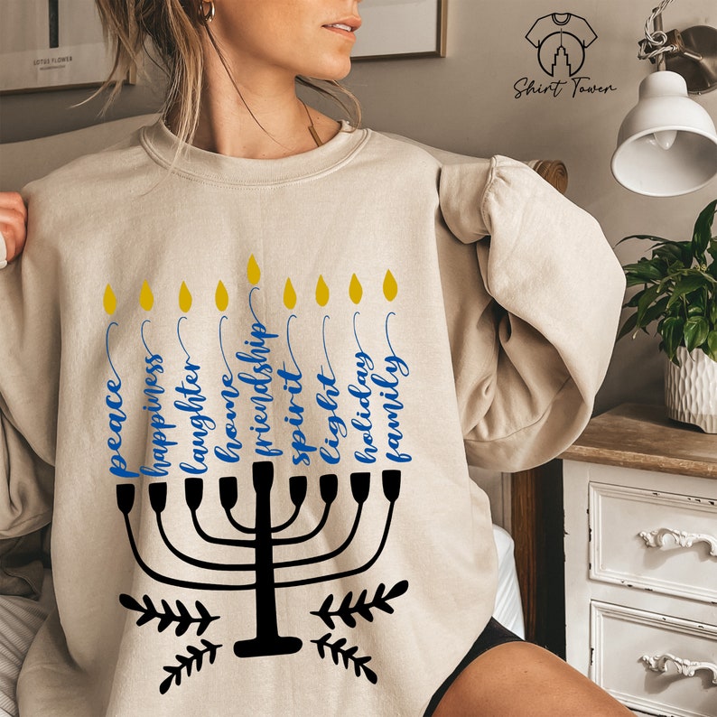 Puede incluir: Una sudadera beige con un dise&ntilde;o de menor&aacute; negro y texto azul. El texto dice "paz", "felicidad", "risa", "hogar", "amistad", "esp&iacute;ritu", "luz", "vacaciones", y "familia".