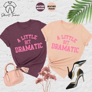 May include: Two t-shirts with the text "A Little Bit Dramatic" in pink lettering. The shirt on the left is heather maroon and the shirt on the right is heather peach.