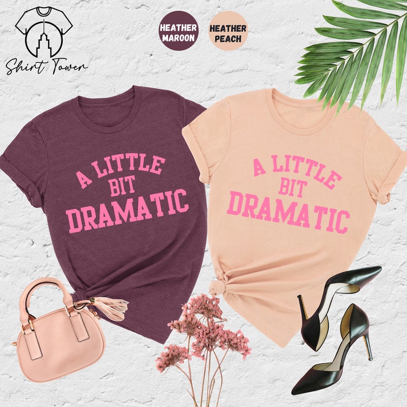 May include: Two t-shirts with the text "A Little Bit Dramatic" in pink lettering. The shirt on the left is heather maroon and the shirt on the right is heather peach.