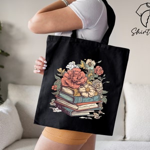 May include: Black tote bag featuring a floral design with a stack of books and flowers. The flowers are pink, white, and yellow. The books are brown, blue, and green.