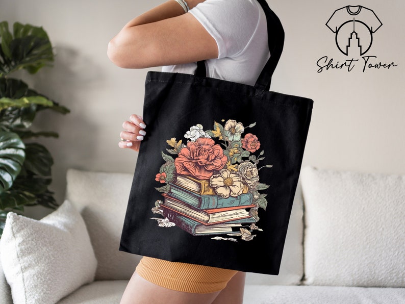 May include: Black tote bag featuring a floral design with a stack of books and flowers. The flowers are pink, white, and yellow. The books are brown, blue, and green.
