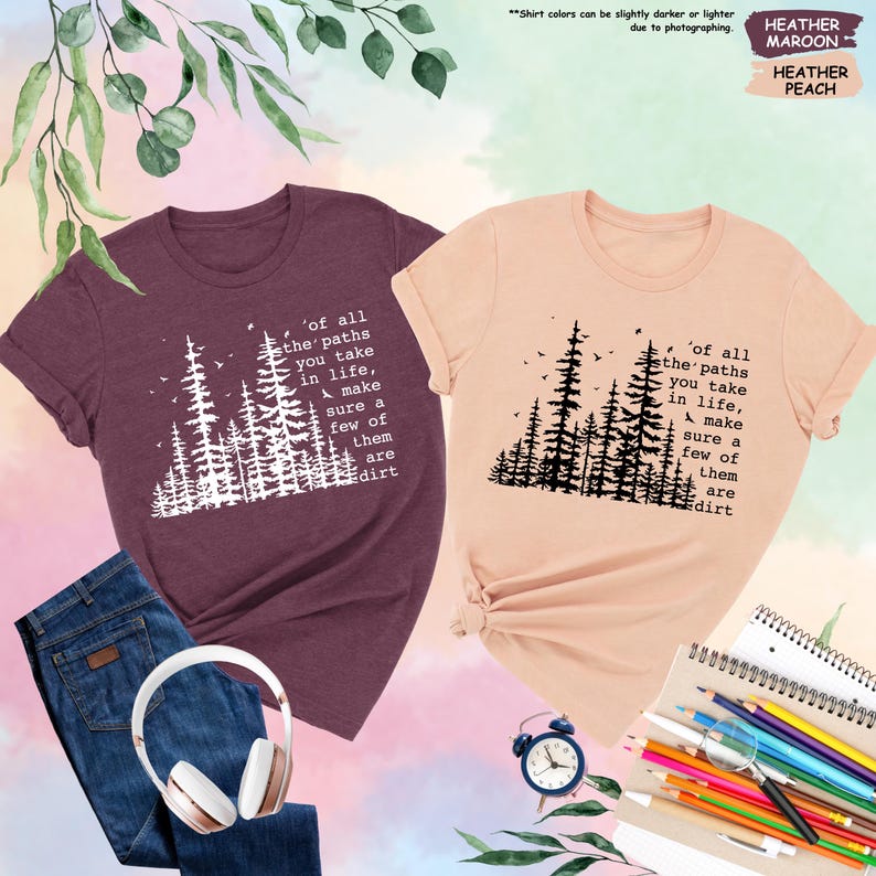 K&ouml;nnte beinhalten: Zwei T-Shirts mit einer Waldszene und dem Text 'of all the paths you take in life, make sure a few of them are dirt'. Das Shirt links ist weinrot und das Shirt rechts ist pfirsichfarben.