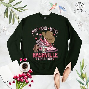 May include: Green sweatshirt with a pink graphic design that says "Boots Booze Besties Nashville Girls Trip". The graphic features a bottle of whiskey, a glass of whiskey, and a pair of pink cowboy boots.