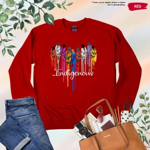 May include: Red long-sleeve shirt with a graphic of a group of people with feathers in their hair, dripping with rainbow colors. The text "Indigenous" is below the graphic.