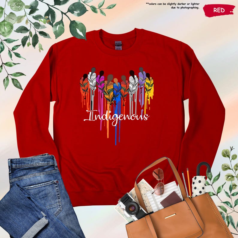 May include: Red long-sleeve shirt with a graphic of a group of people with feathers in their hair, dripping with rainbow colors. The text "Indigenous" is below the graphic.