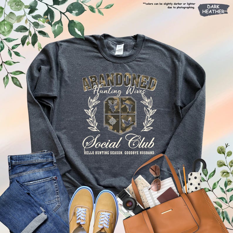 May include: Dark heather gray sweatshirt with the text "ABANDONED Hunting Wives Social Club." The design includes a camouflage emblem and the phrase "HELLO HUNTING SEASON. GOODBYE HUSBAND." The sweatshirt is paired with jeans and tan shoes.