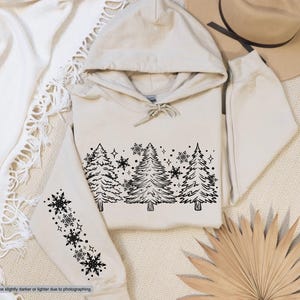 May include: A cream-colored hoodie with a black outline of a winter forest scene on the front. The scene includes pine trees and snowflakes. The hoodie has a black snowflake design on the left sleeve.
