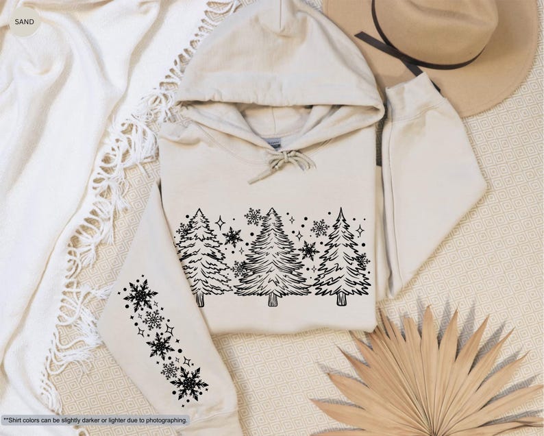 May include: A cream-colored hoodie with a black outline of a winter forest scene on the front. The scene includes pine trees and snowflakes. The hoodie has a black snowflake design on the left sleeve.