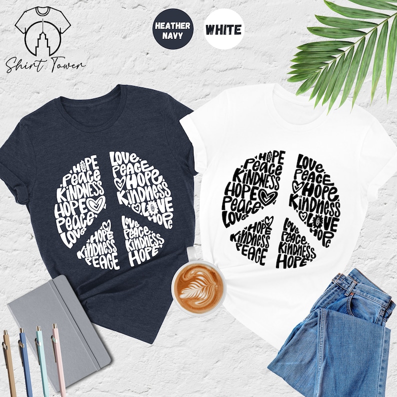May include: Two t-shirts with a peace sign design. The peace sign is made up of the words "Hope", "Peace", "Love", and "Kindness". One shirt is white and the other is heather navy.