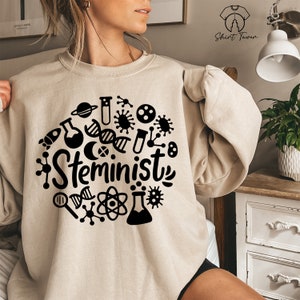 May include: A beige sweatshirt with a black graphic design of a circle with various science-related symbols and the word "Steminist" in the center.