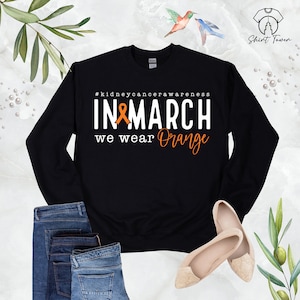 Puede incluir: Sudadera negra con el texto "#kidneycancerawareness IN MARCH we wear Orange" y una cinta naranja.
